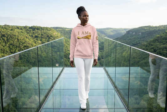 Woman wearing a soft pink Eternal Agape I AM hoodie and white pants while walking across a glass walkway overlooking a green valley.